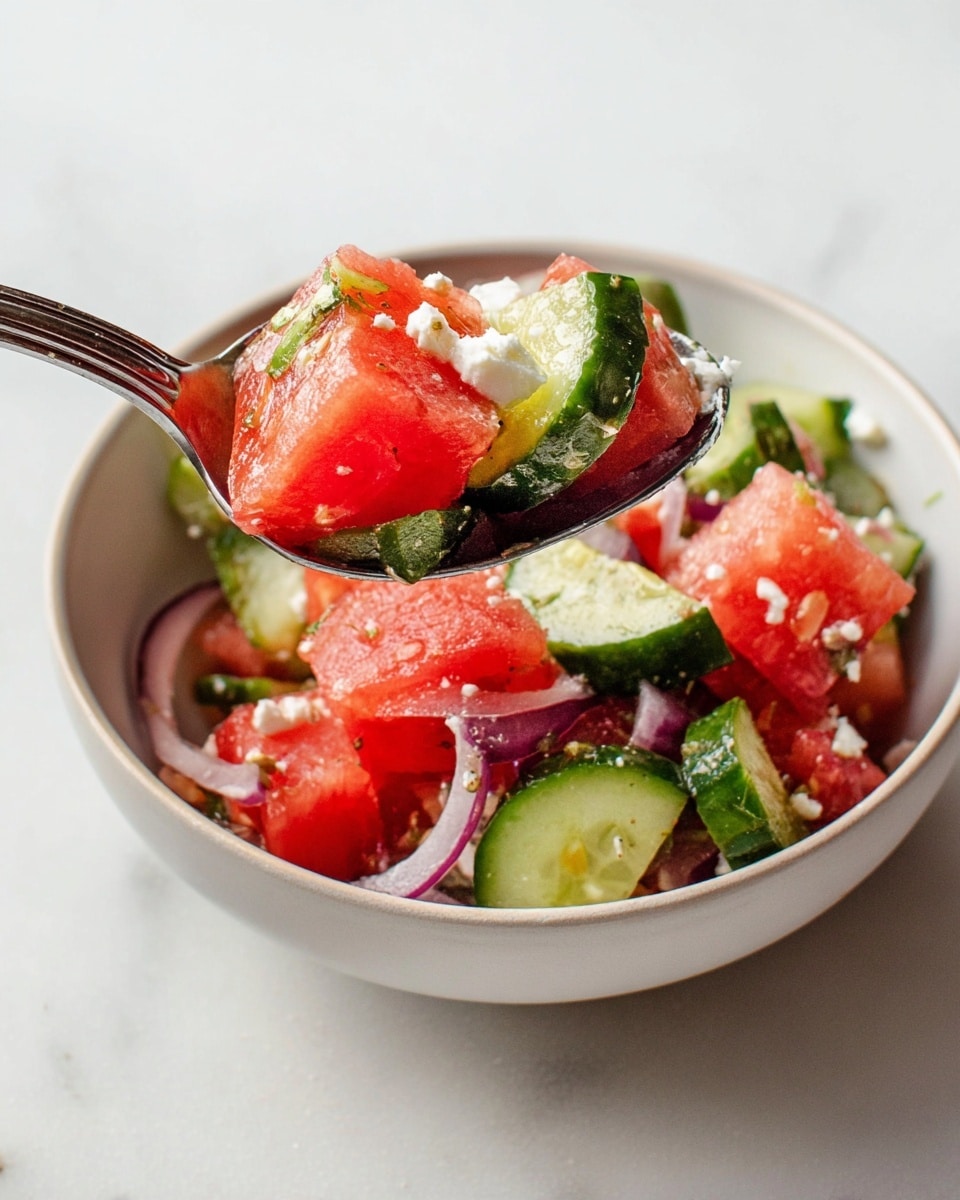 A close-up of a fresh salad in a white bowl showing three main layers: bright red watermelon cubes with a slightly shiny, juicy texture, chopped green cucumber pieces with a light green inside and dark green skin, and small white crumbled cheese scattered evenly throughout. A dark wooden spoon lifts some salad showing the mix clearly. The background is a white marbled texture. Photo taken with an iphone --ar 4:5 --v 7