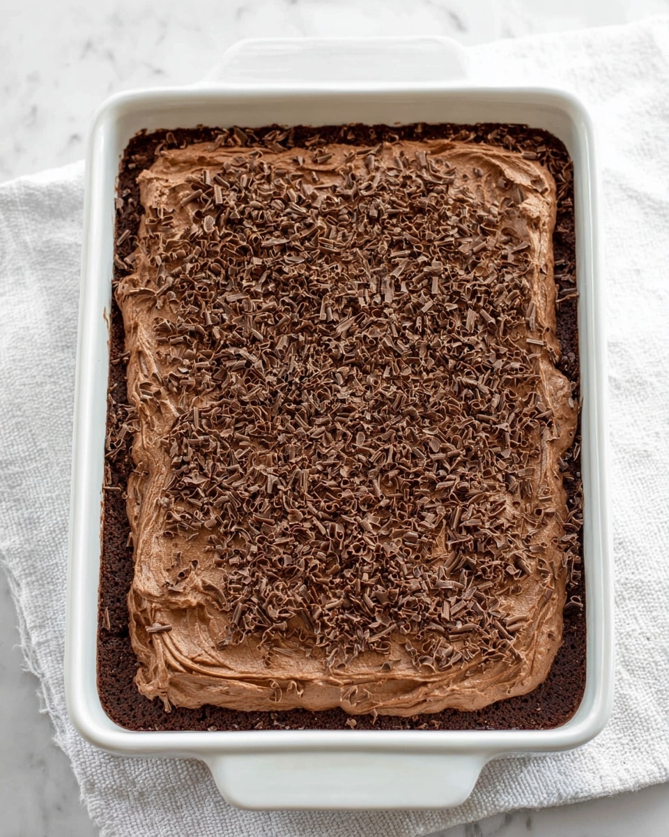 The image shows a rectangle white ceramic baking dish filled with a two-layer chocolate dessert. The bottom layer is a darker brown, dense chocolate cake base visible around the edges. On top is a thick, smooth layer of lighter chocolate mousse spread evenly with soft swirls on the surface. The mousse layer is generously sprinkled with fine, curly chocolate shavings that cover it completely. The whole dish sits on a white marbled surface with a white cloth underneath. Photo taken with an iphone --ar 4:5 --v 7