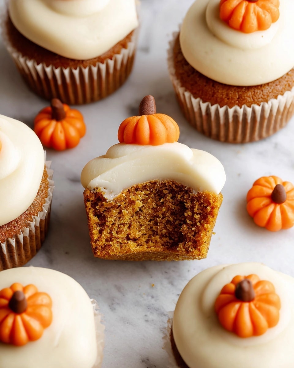 The image shows a close-up of small pumpkin cupcakes on a white marble surface. Each cupcake has a light brown base with a moist texture and is topped with a thick, creamy, off-white frosting shaped in smooth dollops. On top of each frosting dollop, there is a small, detailed orange pumpkin decoration with grooves and tiny brown stems. One cupcake is shown with a bite taken out, revealing the soft crumb inside. The cupcakes are arranged closely together with some extra pumpkin decorations scattered around. Photo taken with an iphone --ar 4:5 --v 7