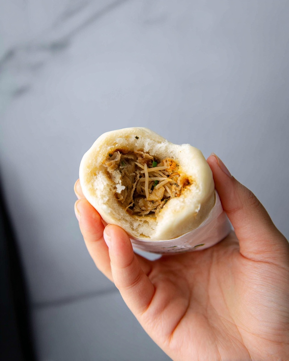 A woman's hand holds a small, beige steamed bun that has a bite taken out of it, revealing its filling. Inside, there is a mix of thin, light brown and beige shredded vegetables or noodles tightly packed within the soft, pale dough. The bun's surface is smooth with slight browning on one side, contrasting with the fluffy, slightly torn white interior dough around the filling. The background is simple with a white marbled texture. Photo taken with an iphone --ar 4:5 --v 7