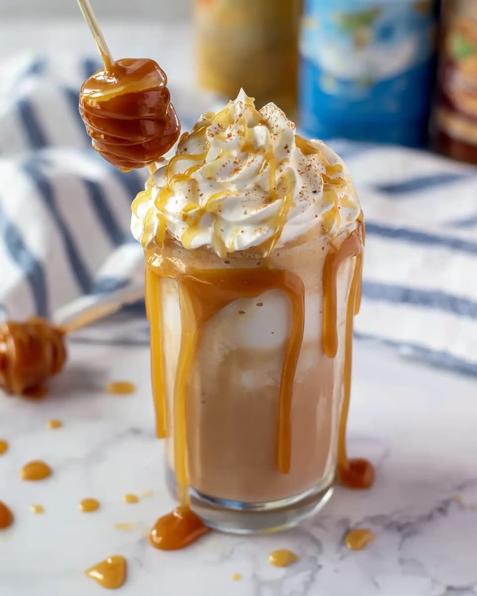 The image shows a glass filled with a layered drink that has a light brown bottom, fading to a lighter cream color at the top. It is topped with a thick, white swirl of whipped cream with light brown specks sprinkled on it. Golden caramel syrup is drizzled over the whipped cream. A caramel-covered lollipop is leaning on the side of the glass. The glass is placed on a white marbled surface with a white cloth with blue stripes in the background and blurred bottles nearby. photo taken with an iphone --ar 4:5 --v 7