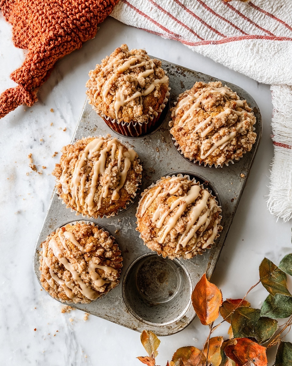 Five muffins sit in a worn metal baking tray with one empty muffin slot. Each muffin has three visible layers: a bottom brown paper liner, a light golden muffin layer, and a crumbly top layer of darker brown streusel sprinkled with crumbs and drizzled with creamy light brown glaze in thick zigzags. The tray rests on a white marbled surface with a white towel that has red stripes and an orange knitted cloth nearby. On the right, some dried orange and green leaves add a natural touch. Photo taken with an iphone --ar 4:5 --v 7