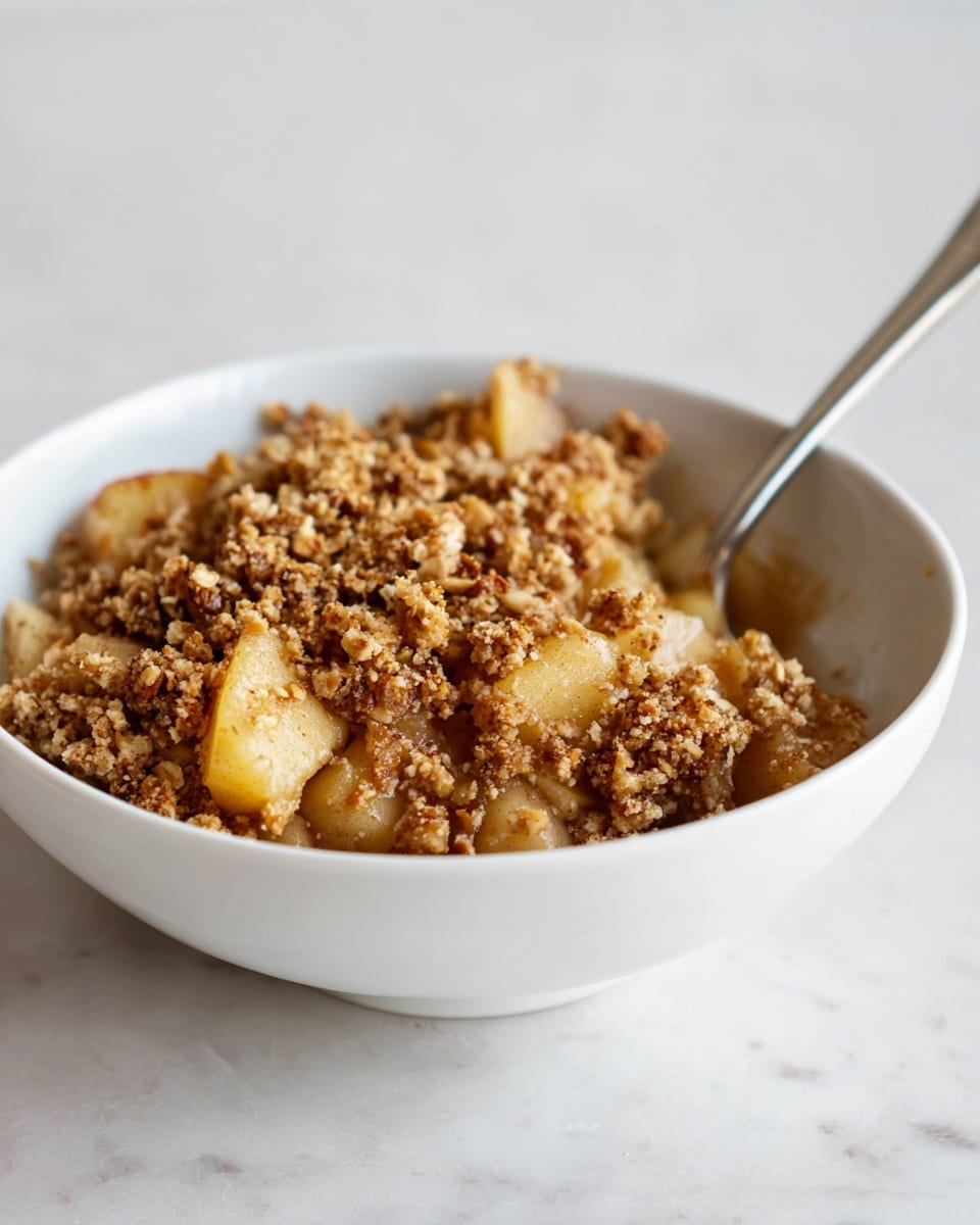 A white bowl filled with a warm apple crumble dessert sits on a white marbled surface. The bottom layer shows soft, light brown cooked apple chunks with a slight golden tint and smooth texture. On top, there is a thick layer of crunchy crumble topping made of small, coarse pieces of oats and nuts in various shades of golden brown, giving a textured and crumbly look. A silver spoon is placed inside the bowl, partially dipped in the dessert, ready to scoop. The light is soft, highlighting the contrast between the tender apples and the crispy topping photo taken with an iphone --ar 4:5 --v 7