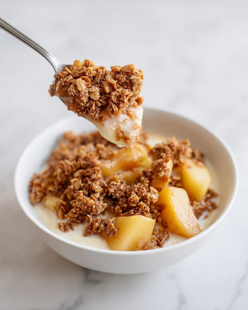 A white bowl filled with a layered dish starting with a thick creamy liquid at the bottom, topped by several clusters of crumbly granola that are golden brown with some darker toasted spots, and mixed with soft, cooked apple chunks that have a light yellow color with a slight orange hue from spices. A spoon is holding a portion of the granola and apple mix above the bowl, showing the texture of the crumbly granola and tender apple pieces clearly. The bowl sits on a white marbled surface. photo taken with an iphone --ar 4:5 --v 7