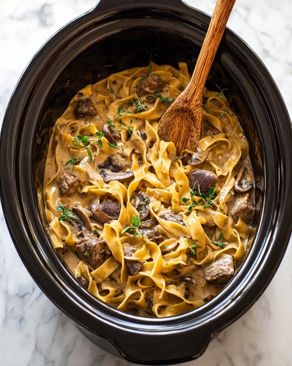 Inside a black slow cooker, there is a creamy dish made of thick, light brown sauce mixed with wide, flat noodles that have a slightly yellow color. The noodles are tangled with slices of dark brown mushrooms and chunks of brown meat. Small green sprigs of herbs are scattered on top adding a touch of freshness. A wooden spoon is partially submerged in the dish, with its handle sticking out upright. The slow cooker sits on a white marbled surface. photo taken with an iphone --ar 4:5 --v 7