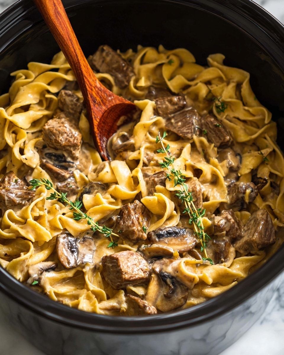 The image shows a close-up view of a creamy pasta dish with three main layers visible: the yellowish wide egg noodles forming the base, the dark brown sliced mushrooms scattered all over, and chunks of tender cooked meat spread evenly within the noodles. Fresh small green sprigs of thyme rest on top, adding a pop of color. The texture of the noodles is soft with a slight gloss from the creamy sauce coating everything, making the dish look rich and hearty. A wooden spoon with a reddish-brown handle is placed inside the pot, stirring the food. The dish is in a black pot sitting on a white marbled surface. Photo taken with an iphone --ar 4:5 --v 7