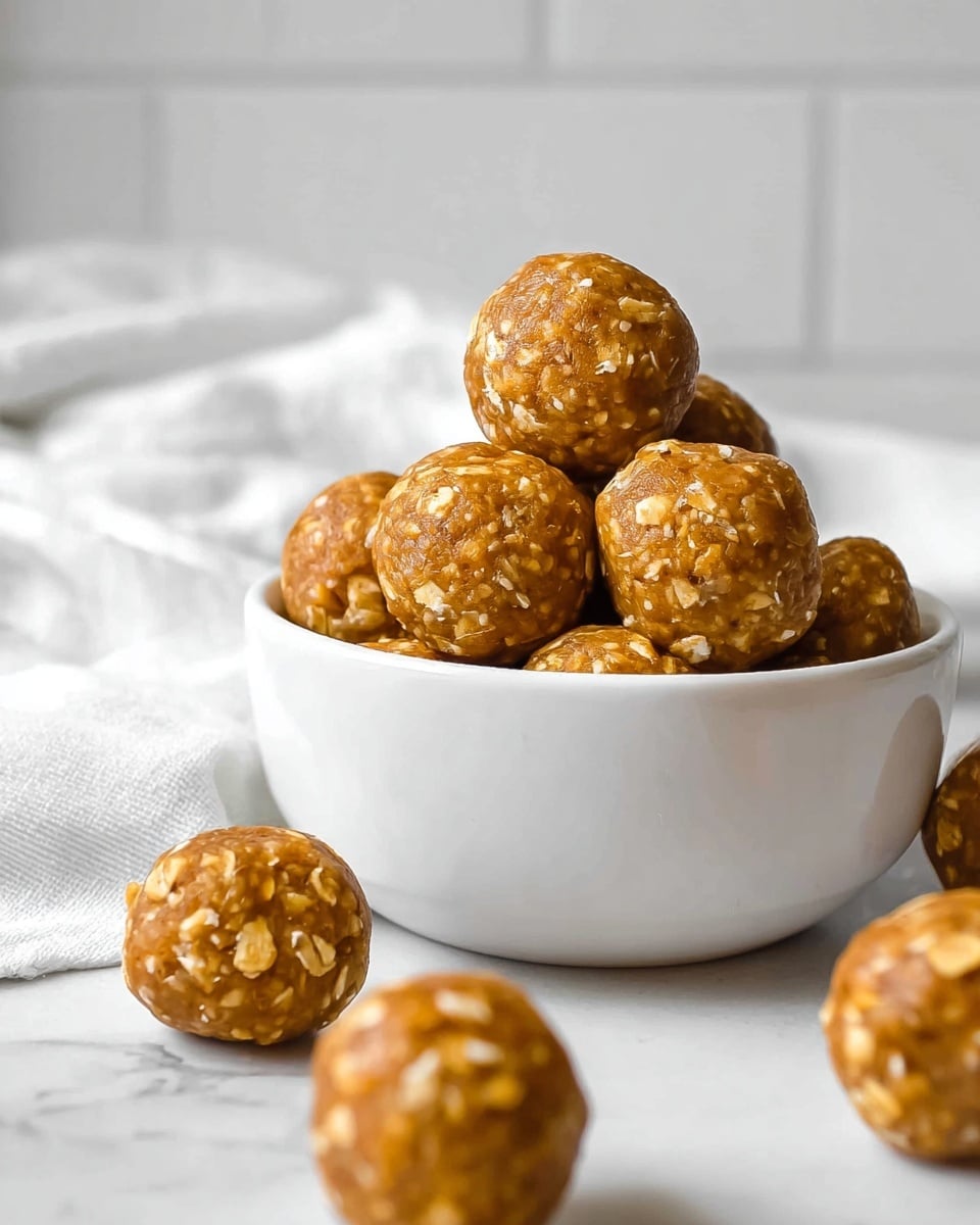 A white ceramic bowl filled with round, golden-brown energy balls showing visible oats texture, stacked and slightly overflowing from the bowl. Several energy balls are scattered around the bowl on a white marbled surface. In the background, a white cloth is softly draped, and white tile walls provide a clean setting. The energy balls have a rough but compact surface with small oat flakes embedded throughout, giving a natural, homemade look. photo taken with an iphone --ar 4:5 --v 7