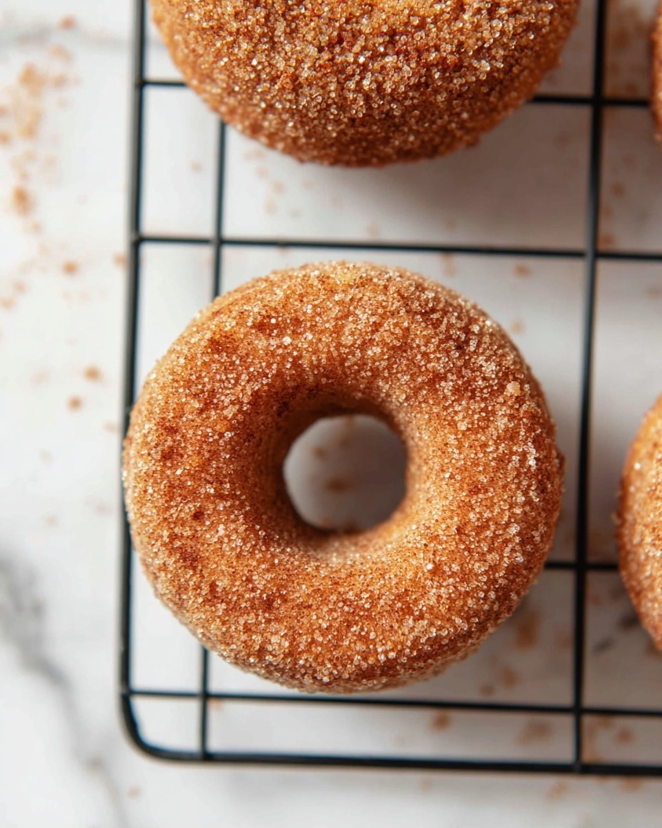The image shows a close-up of a single donut on a black wire cooling rack placed over a white marbled surface. The donut is whole, round, and covered fully with a rough layer of cinnamon sugar crystals, giving it a grainy texture. The donut has a golden-brown color, and the cinnamon sugar coating creates a sparkling effect with small sugar bits scattered around on the surface and rack. The photo is taken from above, highlighting the donut’s symmetrical shape and the hole in the center. Photo taken with an iphone --ar 4:5 --v 7