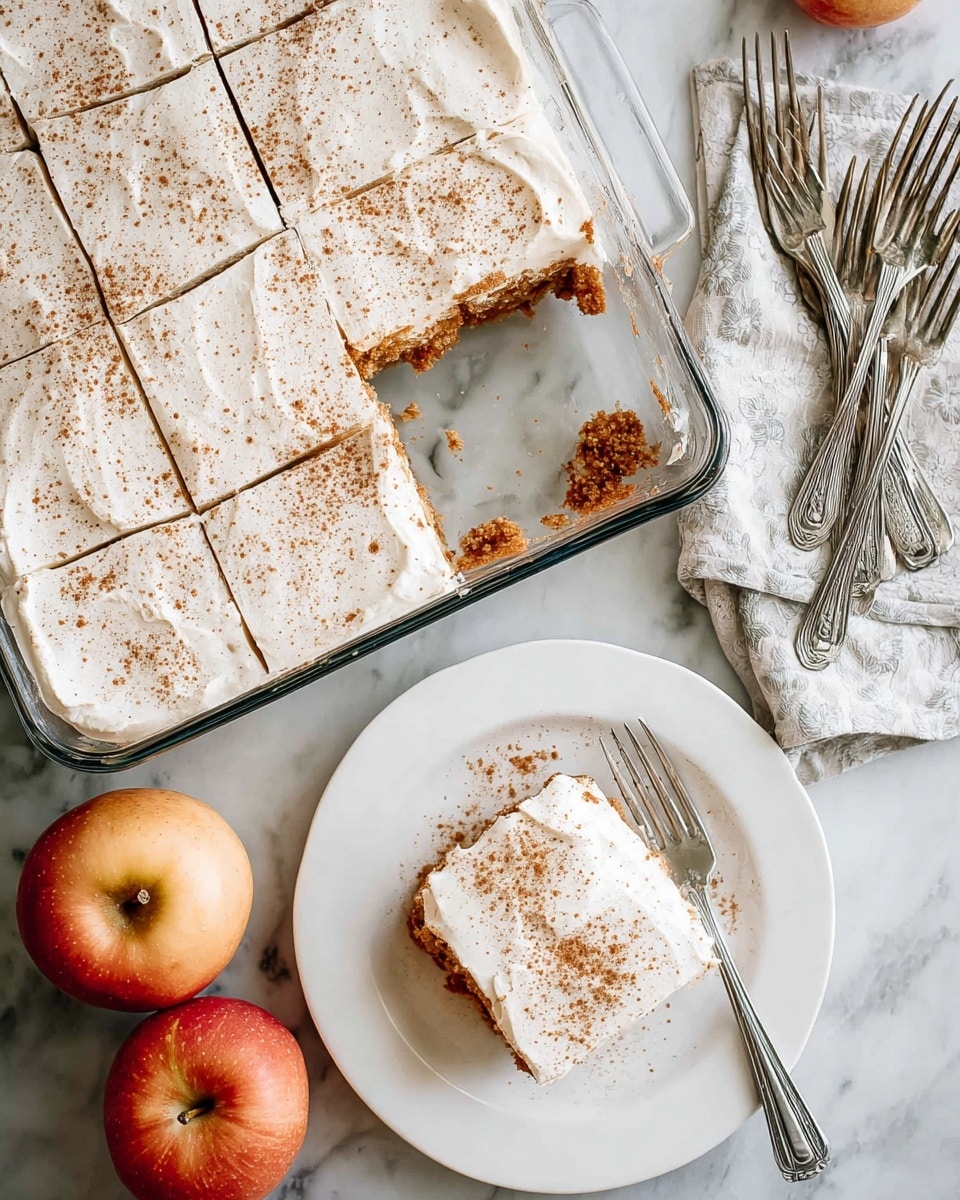 A square glass baking dish contains a dessert cut into square pieces with one piece missing, showing a brown crumbly base layer and a thick, smooth white frosting layer dusted lightly with brown spice on top; next to the dish is a white plate holding one piece of the dessert with the same frosting and spice, accompanied by a silver fork resting on the plate, all placed on a white marbled surface with three apples nearby, the background featuring a few silver forks on a folded cloth. photo taken with an iphone --ar 4:5 --v 7