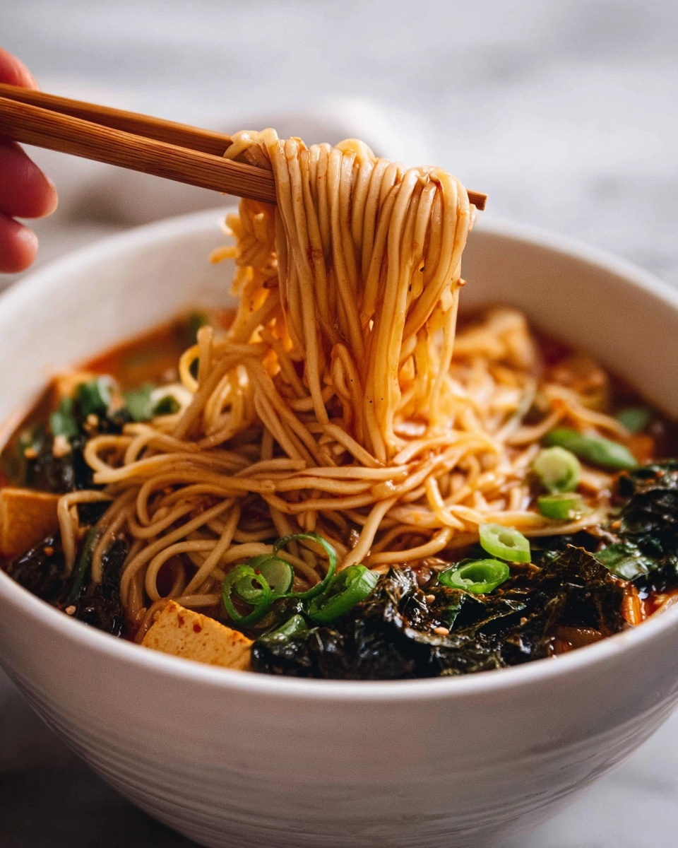 A white bowl filled with a rich broth base and light brown noodles held above it by a pair of wooden chopsticks in a woman's hand. The noodles have a smooth, slightly shiny texture with some green onion slices scattered on top. Around the noodles in the bowl are dark leafy greens and pieces of tofu or a similar ingredient, both soaking in the broth. The background and surface show a white marbled texture. photo taken with an iphone --ar 4:5 --v 7