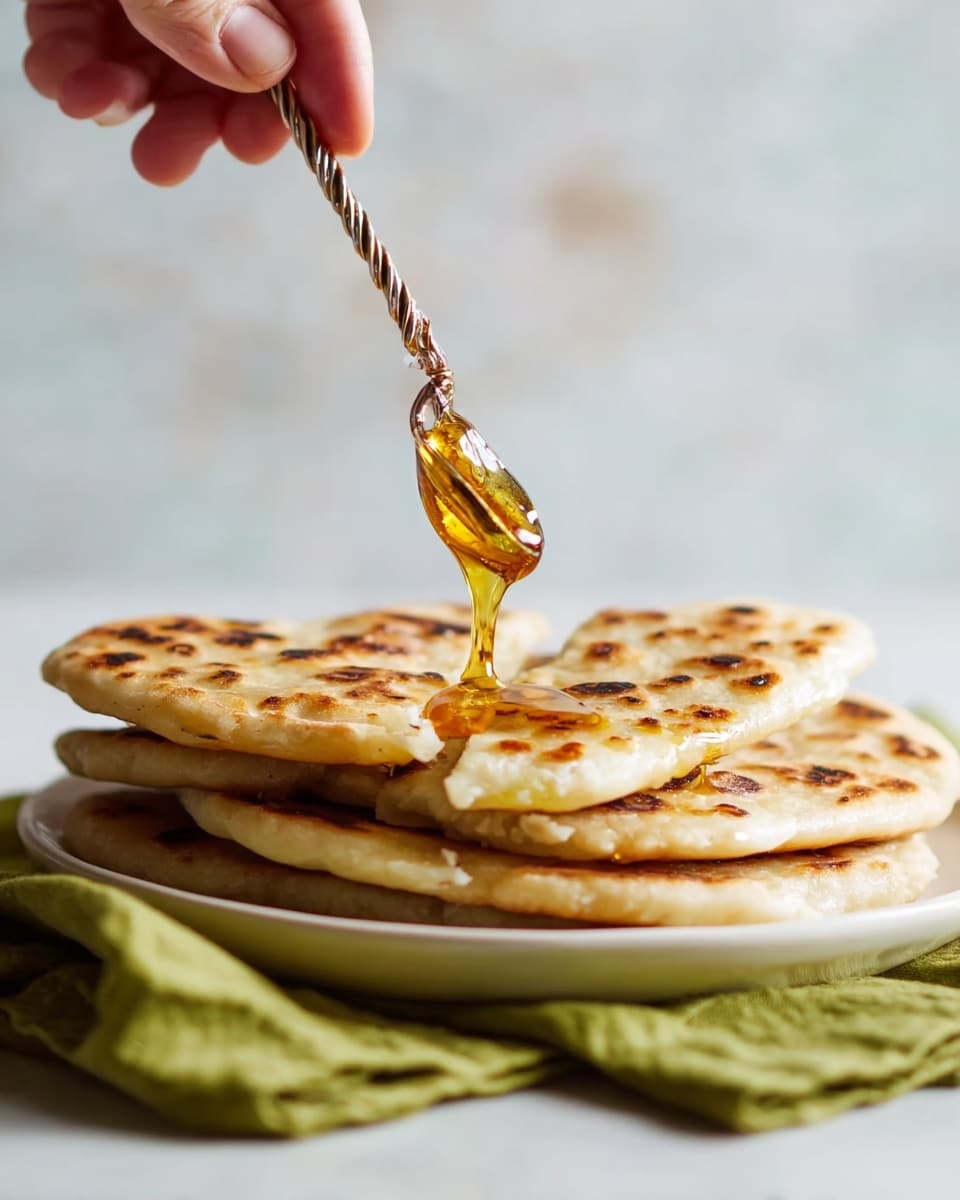 A stack of seven thick flatbreads with light golden brown spots and slightly uneven edges sits on a white plate. Each flatbread layer shows a soft, slightly bumpy texture with some darker toasted patches, creating a warm and inviting look. The stack leans slightly to one side, adding a natural, homemade feel. The plate rests on a green cloth over a woven beige mat, all placed on a white marbled surface. In the background, there is a blurred glass of orange juice. Photo taken with an iphone --ar 4:5 --v 7