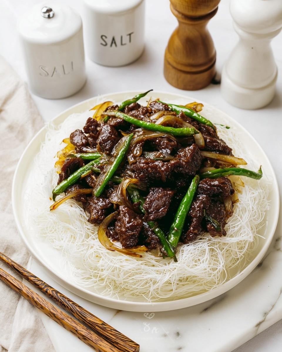 A white plate with a base layer of thin, crispy white noodles arranged in a nest shape, topped with a thick layer of dark brown, glossy cooked meat pieces mixed with sautéed onion slices that are light golden and soft, along with bright green long chili peppers scattered evenly. The dish is placed on a white marbled surface, with a pepper grinder and two white containers labeled