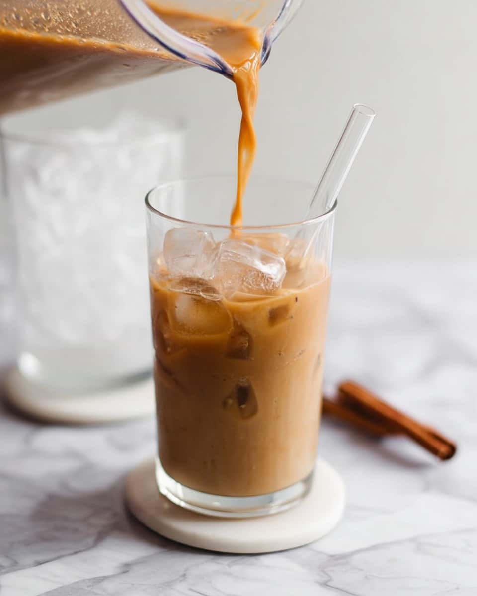 A clear glass filled halfway with light brown iced coffee with visible ice cubes inside, a clear straw standing upright in the glass, more iced coffee being poured from a transparent blender container into the glass, all placed on a white marbled surface with a white coaster underneath, in the background another clear glass filled with ice can be seen slightly out of focus, and a single cinnamon stick lies on the right side on the surface; photo taken with an iphone --ar 4:5 --v 7