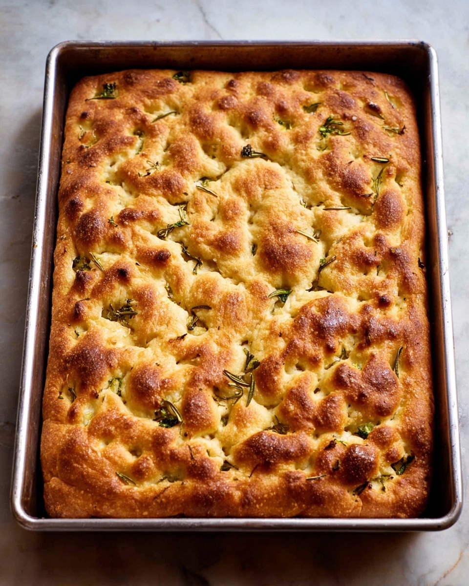 A golden brown focaccia bread with a slightly puffy texture sits evenly spread in a rectangular metal baking pan. The surface shows irregular, rounded bumps and dimples with herbs like rosemary and small green leaves scattered throughout, adding touches of green against the warm, toasted crust. The focaccia has a crisp, slightly uneven top with areas that are darker brown, contrasting the lighter golden parts beneath. The pan rests on a white marbled surface. photo taken with an iphone --ar 4:5 --v 7