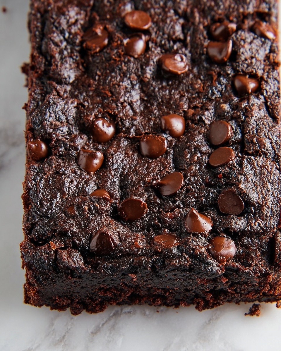 A close-up view of a single-layer dark chocolate cake bar with a rich, deep brown color and a cracked, slightly rough textured top. The surface is generously sprinkled with glossy, dark chocolate chips that are melting slightly in spots, adding shine and smoothness contrast to the rough cake. The cake edges are slightly uneven and dense, showing a moist and fudgy inside beneath the textured dark crust. It sits on a white marbled background. photo taken with an iphone --ar 4:5 --v 7
