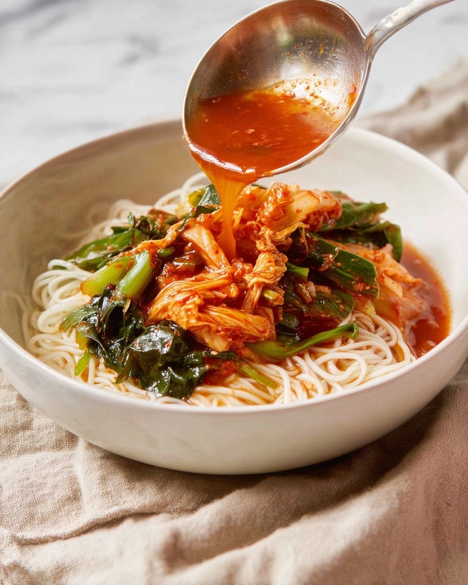 A white bowl holds a base layer of pale, thin noodles arranged loosely. Over the noodles, there are bright green leafy vegetables and large pieces of orange-red kimchi with a slightly wet texture. A silver ladle with some orange-red sauce, bits of green vegetables, and kimchi is positioned above the bowl, pouring more sauce onto the ingredients. The bowl sits on a soft beige cloth, all set against a white marbled surface. Photo taken with an iphone --ar 4:5 --v 7