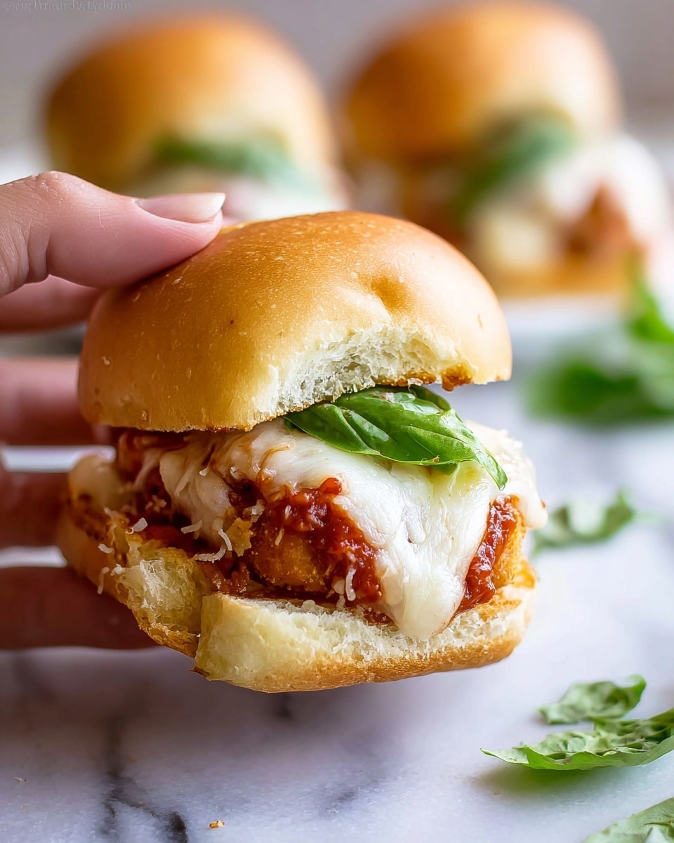 A close-up shows a small sandwich held by a woman's hand, with a soft golden brown bun that looks slightly wrinkled. Inside, there is a main layer of crispy fried item covered with a thick red tomato sauce, topped by melted white cheese that looks creamy and smooth. A fresh bright green basil leaf rests on the cheese. In the blurred background, two more similar sandwiches sit on a white marbled surface, along with some scattered basil leaves. Photo taken with an iphone --ar 4:5 --v 7