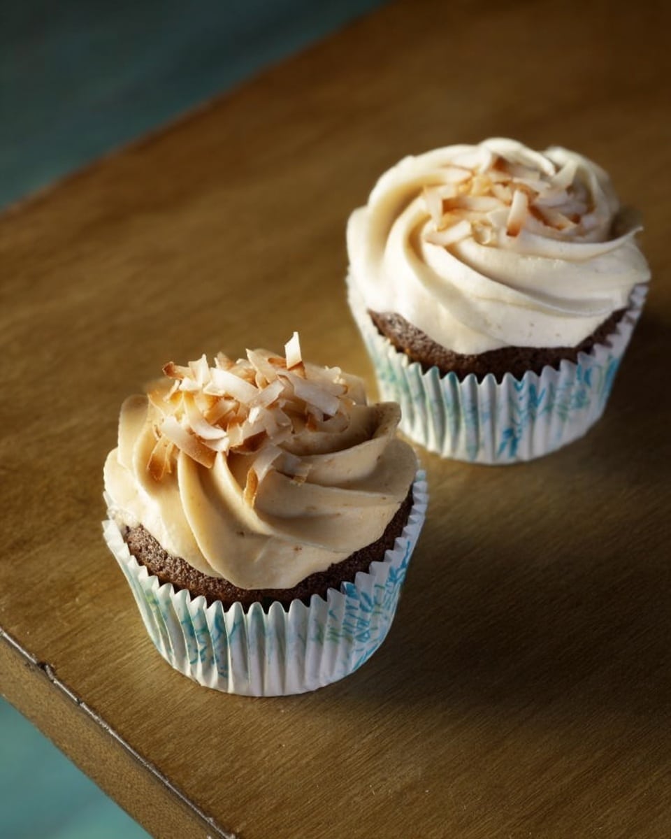 Two chocolate cupcakes sit on a light brown wooden surface. Each cupcake is in a white paper cup with pale blue designs. The cupcakes have a thick layer of light tan frosting swirled on top. One cupcake is topped with toasted coconut slices, adding a slight brown color and rough texture. The other has a small cluster of toasted coconut slices on its smooth frosting. The lighting creates soft shadows around the cupcakes. photo taken with an iphone --ar 4:5 --v 7
