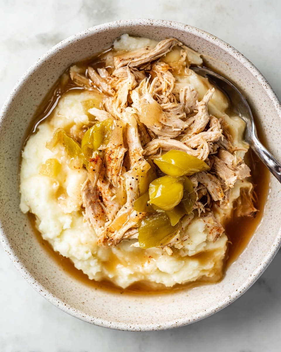 A white speckled bowl holds a dish with two main layers: the bottom layer is smooth, creamy mashed potatoes in a soft white color, topped with shredded cooked chicken that has a light brown, juicy texture and is mixed with a few whole yellow-green peppers. The chicken pieces cover the mashed potatoes unevenly, and brown gravy is visible around the edges, adding shine and moisture to the dish. A silver fork rests on the left side inside the bowl. The bowl is placed on a white marbled surface. photo taken with an iphone --ar 4:5 --v 7