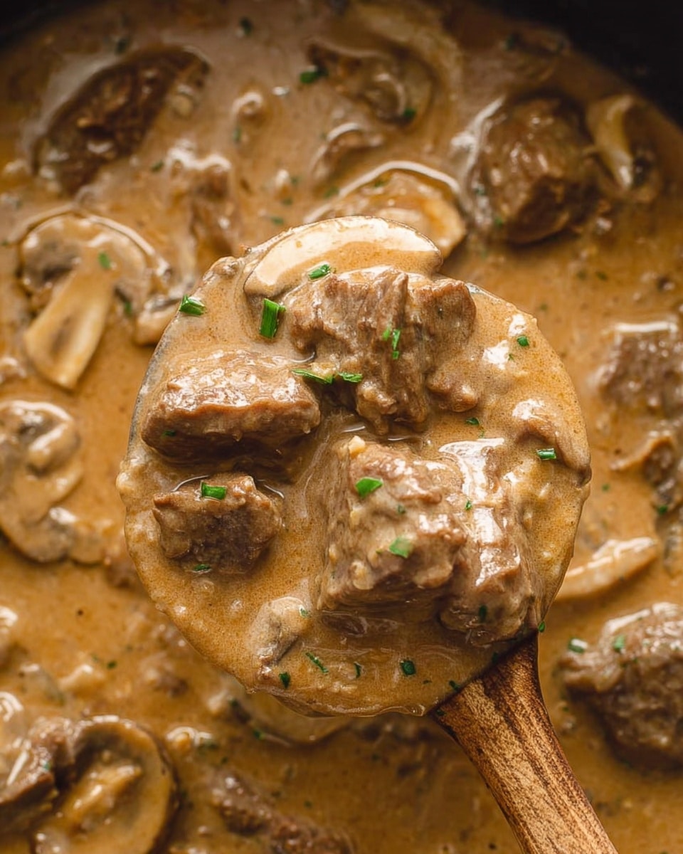 The image shows a white bowl filled with a creamy brown sauce base. Inside the sauce are several dark brown, grilled pieces of meat and light brown mushroom slices scattered throughout. There is a dollop of white mashed potatoes near the center, topped with small green chopped herbs. The bowl sits on a dark textured cloth over a white marbled surface. A silver spoon is placed inside the bowl, angled slightly toward the left side. photo taken with an iphone --ar 4:5 --v 7