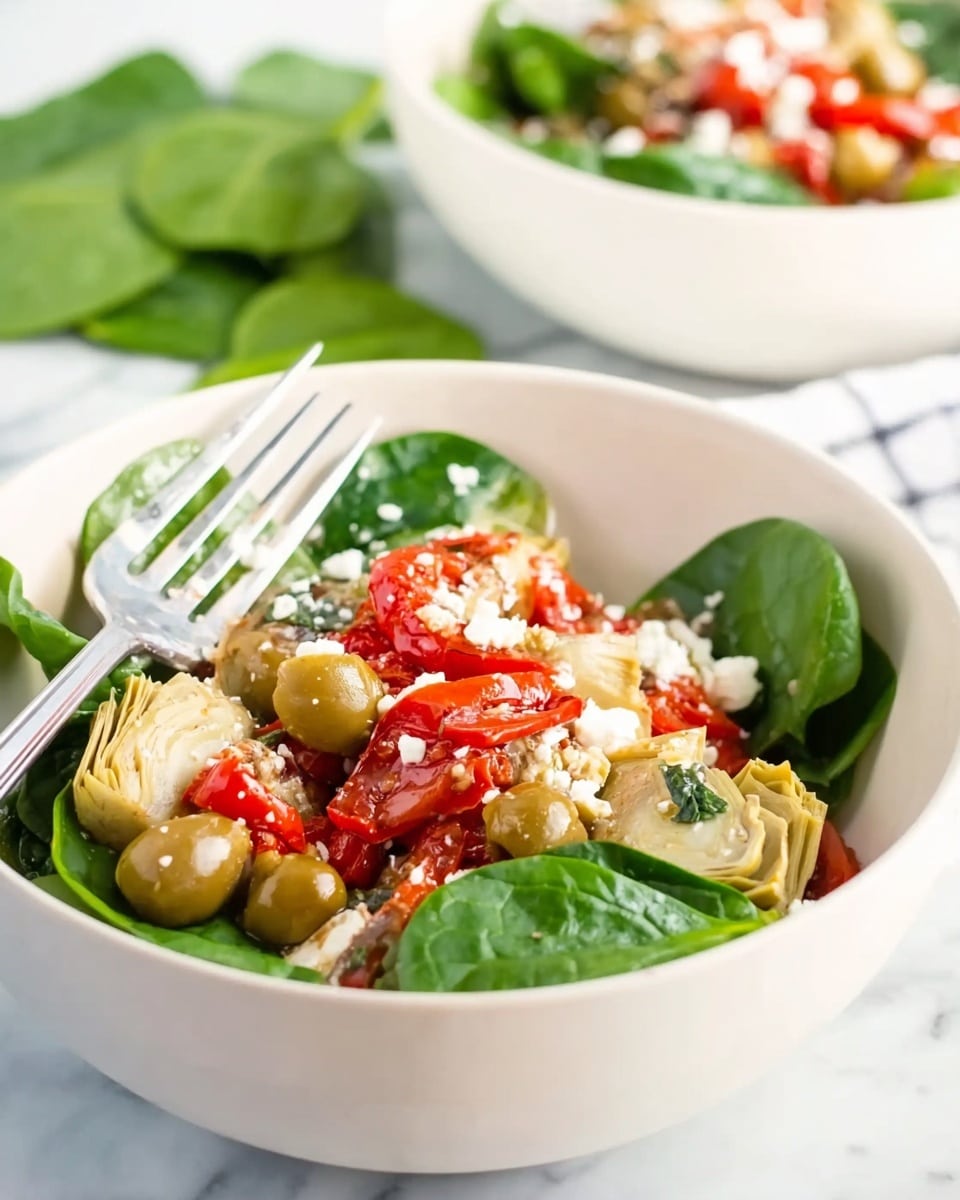 A white bowl filled with a fresh salad showing three clear layers: at the bottom, bright green spinach leaves create a leafy base; the middle layer is made of light brown artichoke hearts and green olives scattered around; the top layer is bright red roasted red pepper strips and small white crumbles of cheese sprinkled evenly. A silver fork rests inside the bowl on the left side and a white marbled surface is beneath the bowl with some green spinach leaves in the background. Another similar bowl is visible in the back right side, slightly out of focus. Photo taken with an iphone --ar 4:5 --v 7