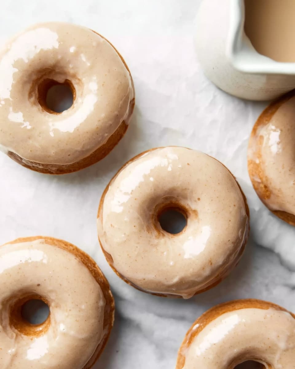 The image shows a group of round donuts with a smooth, light beige glaze on top that has a slightly shiny texture. Each donut has a visible hole in the center. The donuts are placed on a white marbled surface, with some of their edges softly touching each other. In the top right corner, part of a white bowl or cup with a liquid inside is visible. The overall scene has natural lighting that highlights the soft texture of the glaze and the light golden-brown color of the donuts underneath. photo taken with an iphone --ar 4:5 --v 7