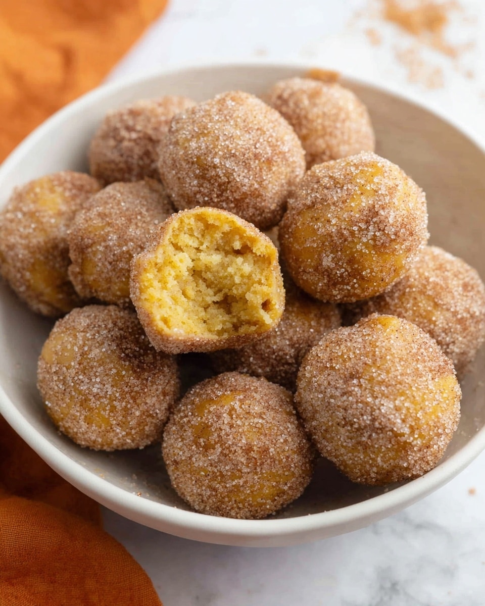 A white oval plate with a thin gold rim holds twenty small round balls arranged closely together in a loose triangular shape. Each ball is golden-brown and coated evenly with a grainy layer of cinnamon sugar, giving a slightly rough texture. Around the plate, three cinnamon sticks and a larger cinnamon piece lie on the white marbled surface. The top left corner shows a blurred bunch of orange reeds in a white vase, with a soft orange and white striped cloth partially visible at the bottom left of the image. The lighting is soft and warm, highlighting the texture and color of the balls. photo taken with an iphone --ar 4:5 --v 7