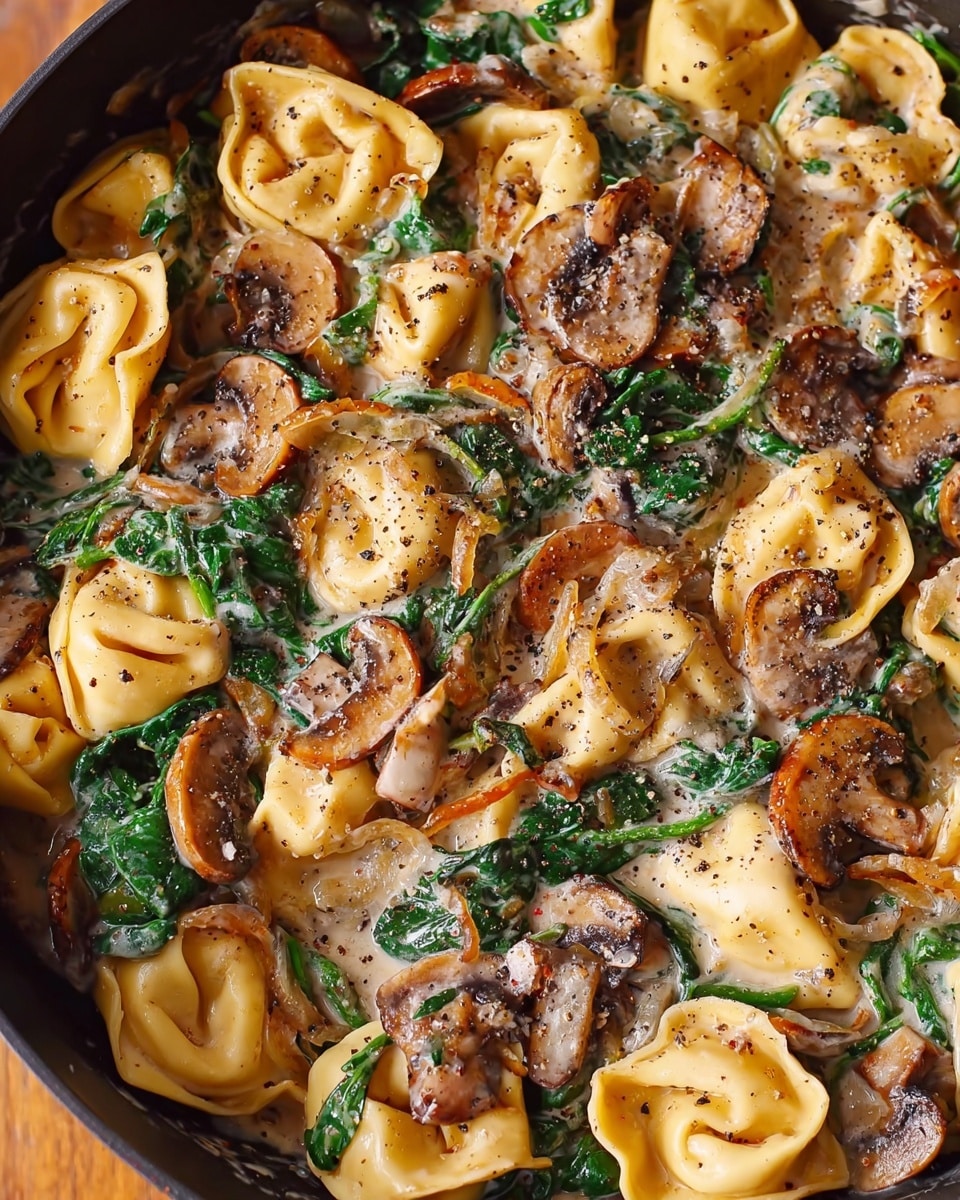 Creamy Spinach and Mushroom Tortellini with Caramelized Onions Recipe 5 The image shows a close-up of a pan filled with soft yellow tortellini pasta mixed with sautéed mushrooms that have a brown and creamy texture, wilted dark green spinach leaves, and bits of light brown caramelized onions. The dish is covered in a creamy white sauce with specks of black pepper sprinkled all over, giving the meal a seasoned look. The pan sits on a wooden surface, but the main focus is the creamy, colorful, and textured mix inside the pan. photo taken with an iphone --ar 4:5 --v 7