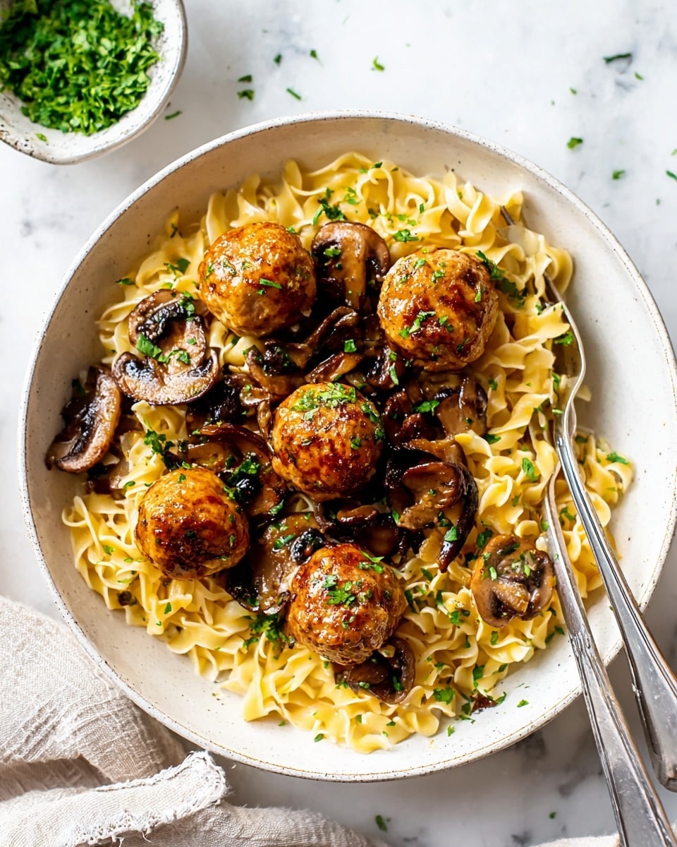 A white bowl on a white marbled surface holds a serving of light yellow egg noodles as the bottom layer, topped with five round, golden-brown meatballs evenly placed. Scattered around and on the meatballs are sautéed brown mushrooms with a glossy texture, sprinkled with fresh green chopped herbs. A silver fork rests inside the bowl, partly touching the noodles and mushrooms. In the background, there is a small bowl filled with more green herbs. Photo taken with an iphone --ar 4:5 --v 7