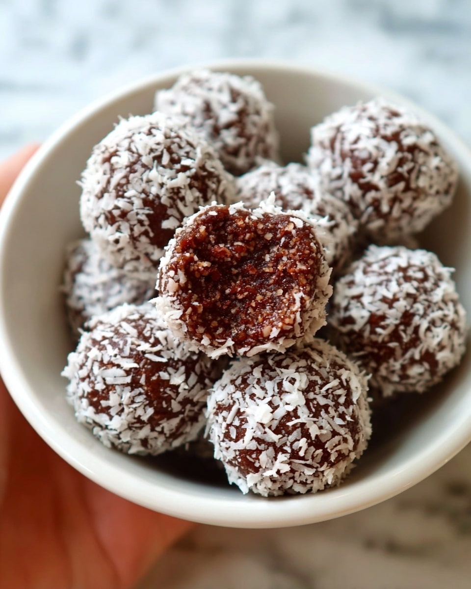 Chocolate Coconut Date Balls (No Bake) (Vegan+GF) Recipe 6 A woman's hand is holding a dark brown round ball with a rough texture, coated with white shredded coconut flakes. The ball is above a white plate filled with a thick layer of white shredded coconut, spread in a circular shape. More dark brown balls with a similar texture are scattered on the upper part of the plate. The plate is placed on a white marbled surface. photo taken with an iphone --ar 4:5 --v 7