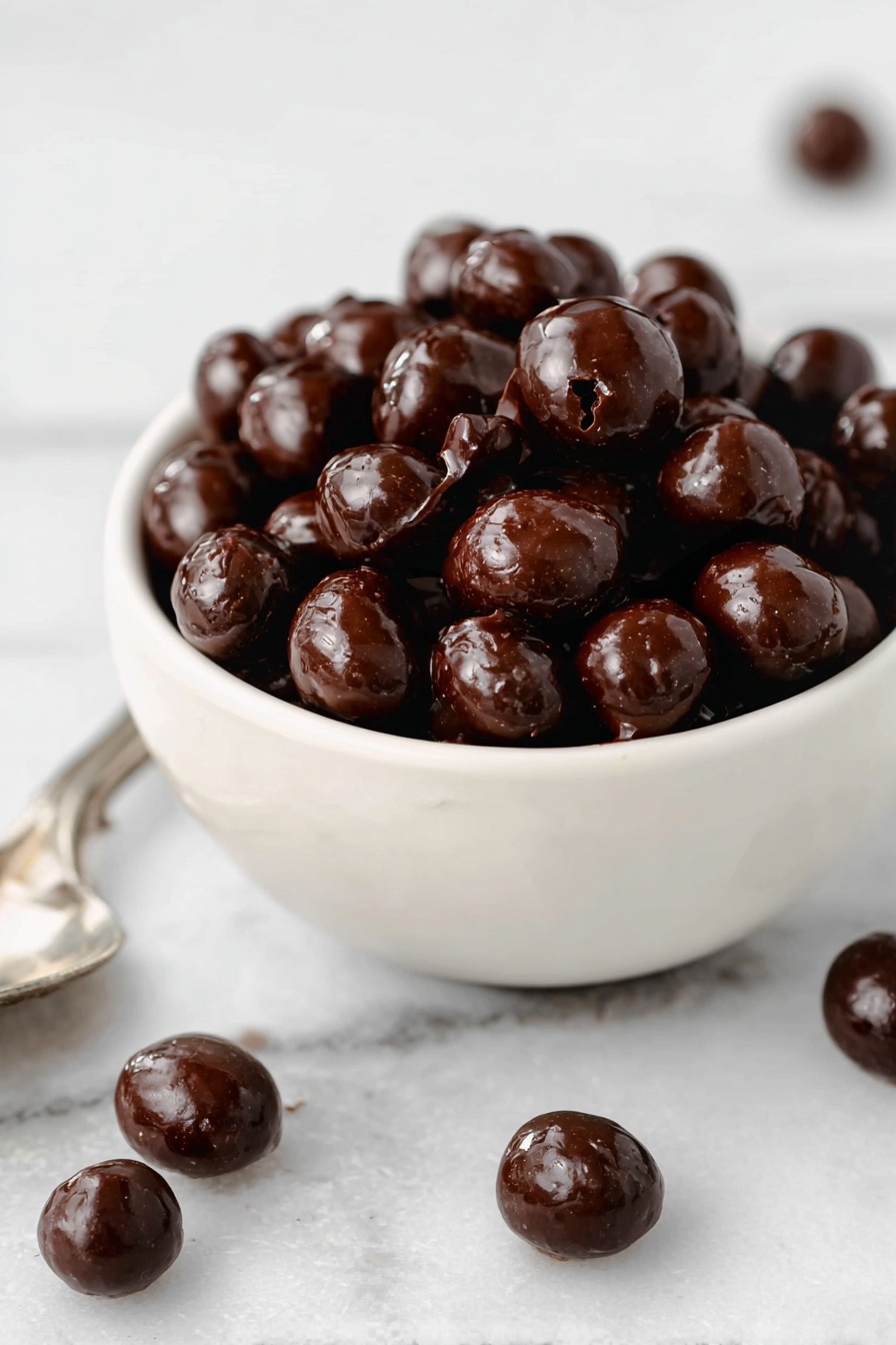 A small white bowl filled with shiny, dark brown chocolate-coated nuts piled high inside. The nuts have a smooth and glossy texture with some cracks showing their natural shape. Around the bowl on a white marbled surface, a few more chocolate-coated nuts are scattered, with some resting on a silver spoon at the bottom left corner. Photo taken with an iphone --ar 2:3 --v 7 - Chocolate Covered Espresso Beans, espresso bean confection, coffee chocolate snacks, caffeinated treat recipe, homemade espresso candies