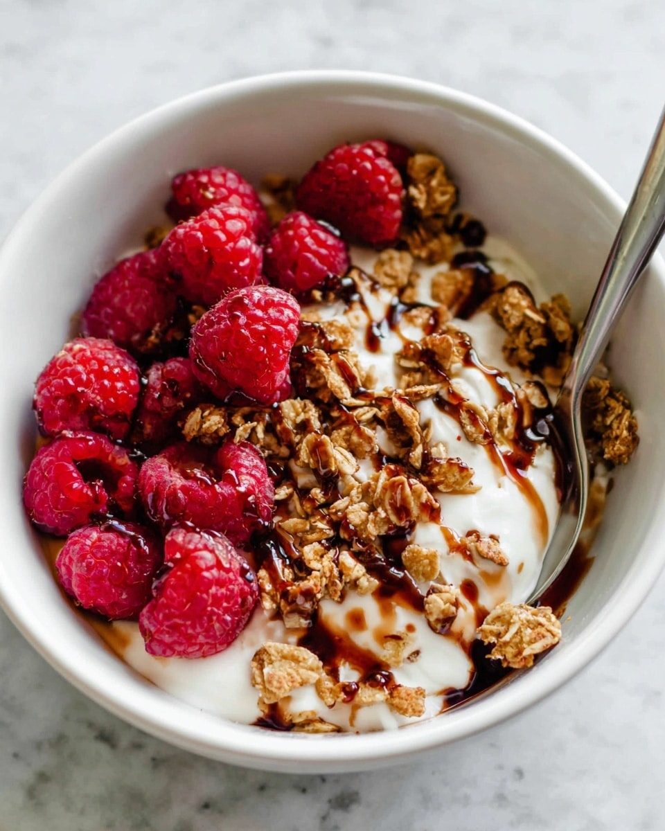 The dish is shown in a white bowl filled with three main layers. The bottom layer is smooth white yogurt. On top of the yogurt is a sprinkle of light brown granola clusters, which have a crunchy texture. Scattered among the granola are bright red raspberries, round and plump with a bumpy surface. Over the top, there are streaks of dark brown syrup drizzled over the yogurt, granola, and raspberries, adding a glossy look. A metal spoon is partly inside the bowl, with its handle leaning out. The bowl sits on a white marbled surface. photo taken with an iphone --ar 4:5 --v 7