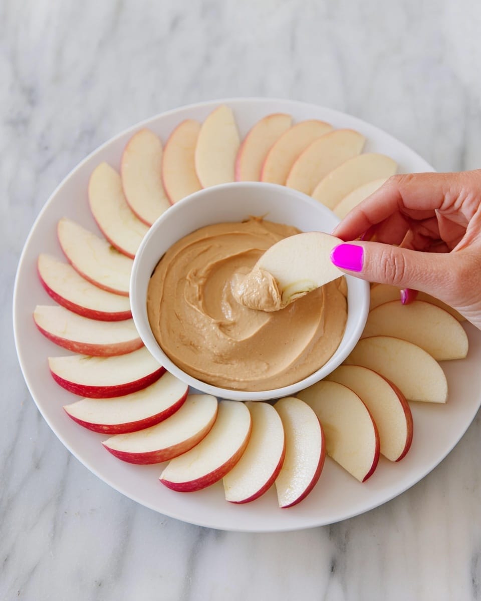 3-Ingredient Cream Cheese Apple Dip Recipe 5 A white plate holds a white bowl filled with a smooth, light brown creamy dip. Around the bowl, there is a single layer of apple slices, each slice pale cream with light red edges, arranged in a neat circle. A woman's hand with bright pink nail polish is lifting an apple slice, ready to dip it into the creamy spread. The plate sits on a white marbled texture surface. photo taken with an iphone --ar 4:5 --v 7