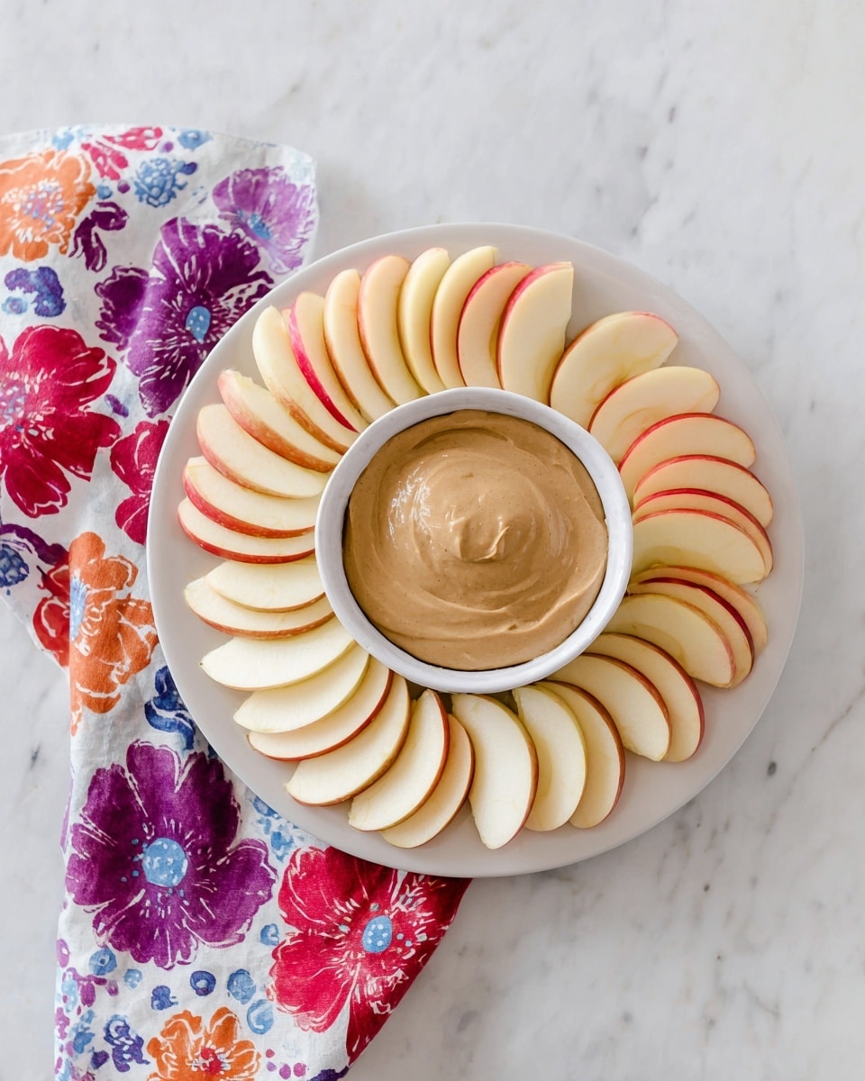 3-Ingredient Cream Cheese Apple Dip Recipe 6 A white plate is filled with two layers of apple slices arranged in a circular pattern. The apple slices on the outside have a pale cream color with red edges, and the inner layer of slices shows darker red skin with a pale side. In the center of the plate, a white bowl holds a thick, smooth, light brown dip with soft swirls on the surface. The plate is set on a white marbled surface, and a colorful cloth with blue, black, and red spots is partially visible on the left side. photo taken with an iphone --ar 4:5 --v 7