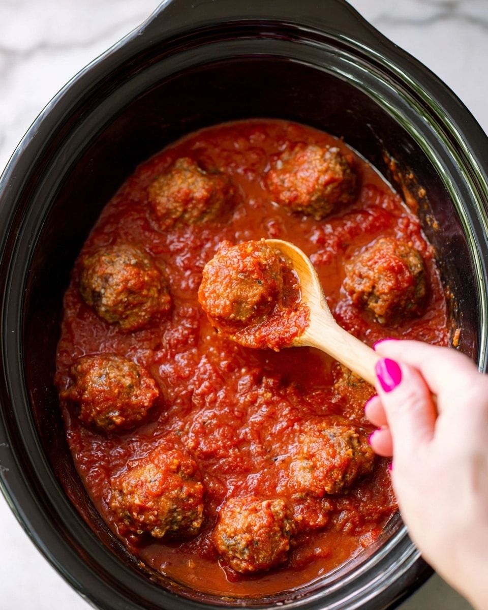Slow Cooker Italian Meatballs Recipe 5 The image shows a black slow cooker filled with a thick, textured red tomato sauce covering nine round, brown meatballs. A woman's hand with pink painted nails is holding a wooden spoon that lifts one meatball, showing its moist, uneven surface coated in sauce. The red sauce has bits of herbs and a slightly chunky texture, spreading evenly inside the slow cooker. The background is a white marbled surface. photo taken with an iphone --ar 4:5 --v 7