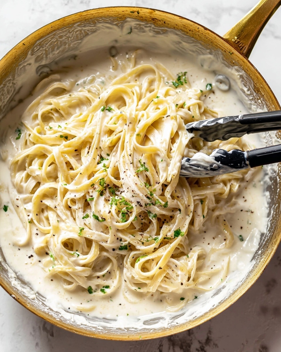 A close-up view of one big silver pan with a golden handle, filled with creamy white sauce and fettuccine pasta mixed in it. The pasta is pale yellow, covered in thick smooth white cream, sprinkled with small green herbs and black pepper dots. A pair of black kitchen tongs is holding some pasta in the middle right of the pan, showing the sauce clinging to the pasta strands. The pan sits on a white marbled surface. Photo taken with an iphone --ar 4:5 --v 7