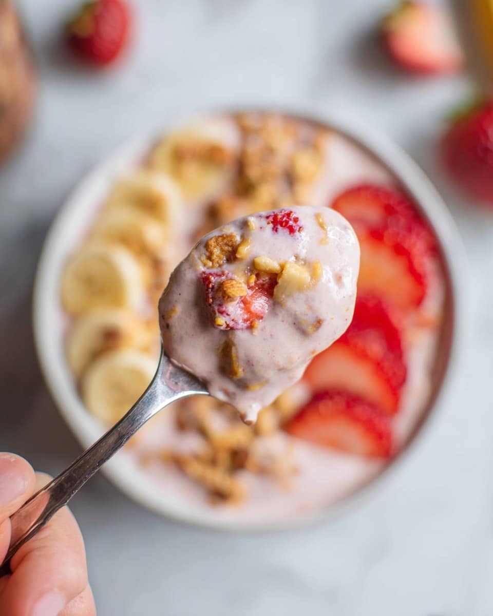 A close-up image of a metal spoon held by a woman's hand, filled with a light pink creamy mixture containing small pieces of red strawberries, tan nut chunks, and small bits of granola. In the blurred background, a white bowl with a similar pink base mixture can be seen, layered with bright red strawberry slices on one side, light beige banana slices on another, and sprinkled with golden brown granola on top. The whole scene is set on a white marbled surface. photo taken with an iphone --ar 4:5 --v 7