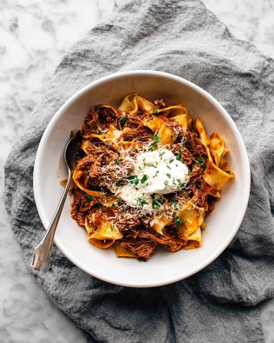 A white bowl holds a serving of wide, flat pasta ribbons in a rich brown-red sauce with shredded meat mixed throughout. On top, there is a dollop of creamy white cheese or cream, sprinkled with grated cheese and chopped green herbs. A silver spoon rests in the bowl on the left side. The bowl is placed on a wrinkled gray cloth over a white marbled surface. photo taken with an iphone --ar 4:5 --v 7