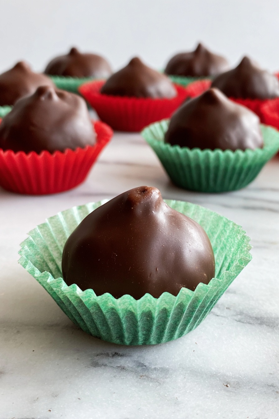 A group of small round chocolate-covered treats sits on a white marbled surface, each resting in a crinkled paper cup. The closest treat is in a green paper cup, showing a smooth, shiny dark brown chocolate coating with a small peak at the top. Behind it, several more chocolates are in red and green paper cups, all similar in shape and texture, creating a neat cluster. The soft light highlights the glossy finish of the chocolates against the white marbled background. photo taken with an iphone --ar 2:3 --v 7 - Chocolate Covered Cherries, Chocolate Covered Cherries recipe, how to make chocolate covered cherries, homemade chocolate cherries, easy chocolate dipped cherries