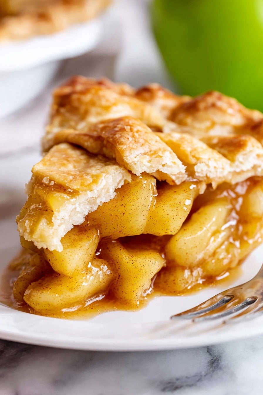 Classic Homemade Apple Pie Recipe 6 A close-up of a slice of apple pie showing a flaky, golden-brown top crust with uneven, rustic edges. Beneath this crust, the filling has soft, light yellow apple chunks coated in a shiny, amber-colored sauce with specks of cinnamon. The pie rests on a white plate placed on a white marbled surface. In the background, there is a blurred green apple. A shiny silver fork is partially visible at the bottom right corner of the image. photo taken with an iphone --ar 2:3 --v 7 - Classic Homemade Apple Pie, apple pie recipe, homemade apple pie, flaky pie crust, best apple pie tips