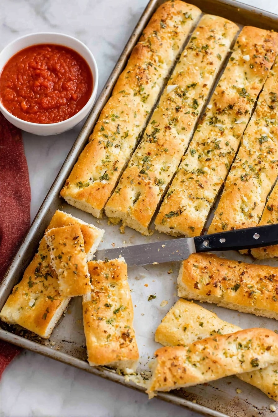 Copycat Pizza Hut Breadsticks Recipe 8 The image shows a metal baking tray filled with rectangular, golden-brown breadsticks, each sprinkled with green herbs and bits of garlic on top. The breadsticks are arranged in three rows inside the tray, and a few pieces are placed outside the tray on a white marbled surface. A knife with a black handle rests on the breadsticks inside the tray. Next to the tray, there is a white bowl filled with red marinara sauce, with one breadstick partially dipped in it. The breadsticks look soft inside and slightly crispy on the outside. Photo taken with an iphone --ar 2:3 --v 7 - Copycat Pizza Hut Breadsticks, homemade pizza breadsticks, soft buttery garlic breadsticks, how to make Pizza Hut breadsticks, best homemade breadsticks