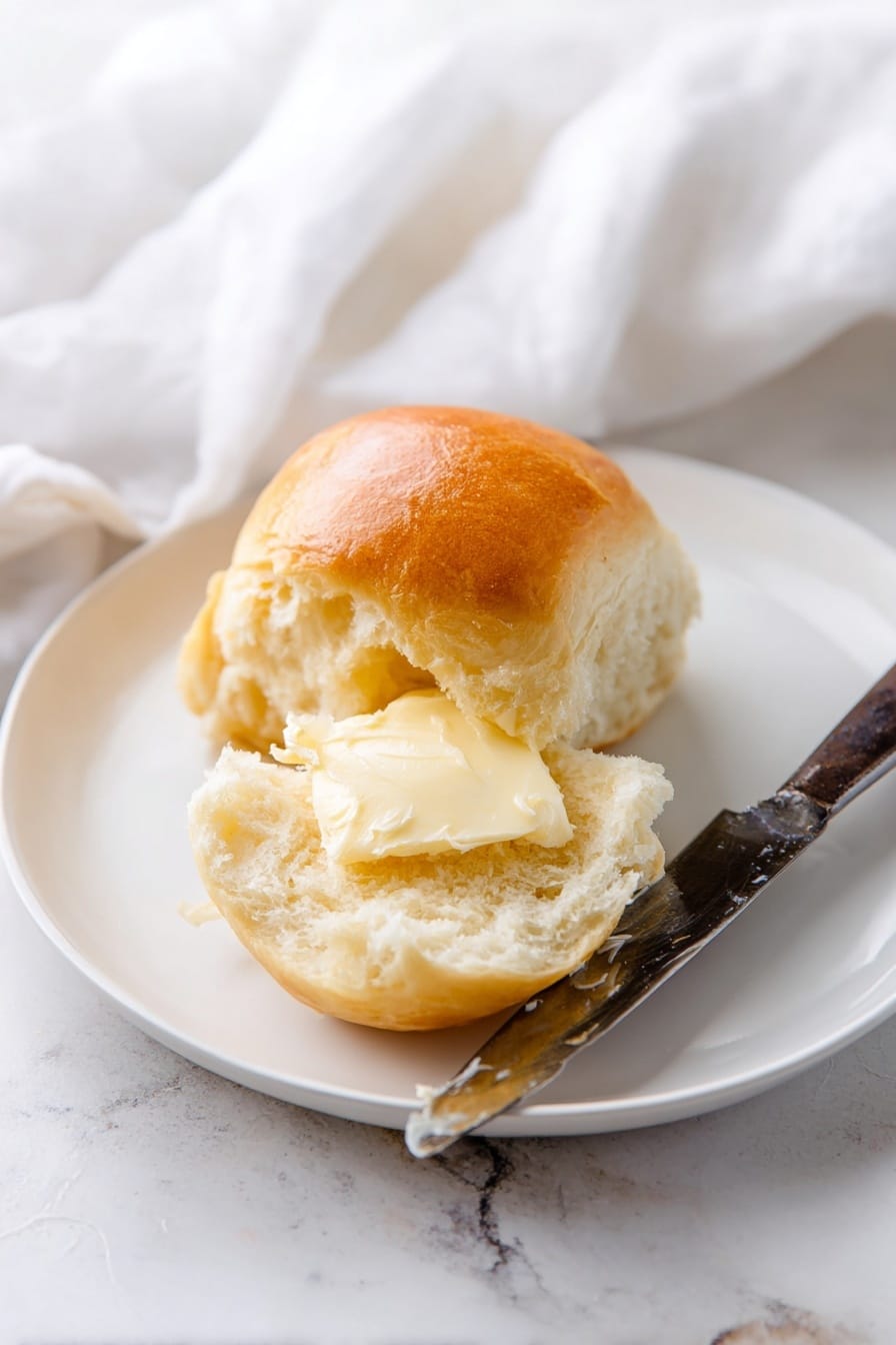Soft & Fluffy Dinner Rolls Recipe 8 A white plate on a white marbled surface holds a soft, golden brown bread roll that is split open. The inside of the roll is light and fluffy with a smooth, shiny spread of butter melted into the upper half. Beside the roll lies an old-fashioned knife with a dark handle and a thick, creamy layer of butter on its blade. In the background, a white cloth is softly draped, adding a cozy feel to the scene. photo taken with an iphone --ar 2:3 --v 7 - Soft & Fluffy Dinner Rolls, dinner rolls recipe, easy dinner roll recipe, homemade dinner rolls, soft bread rolls