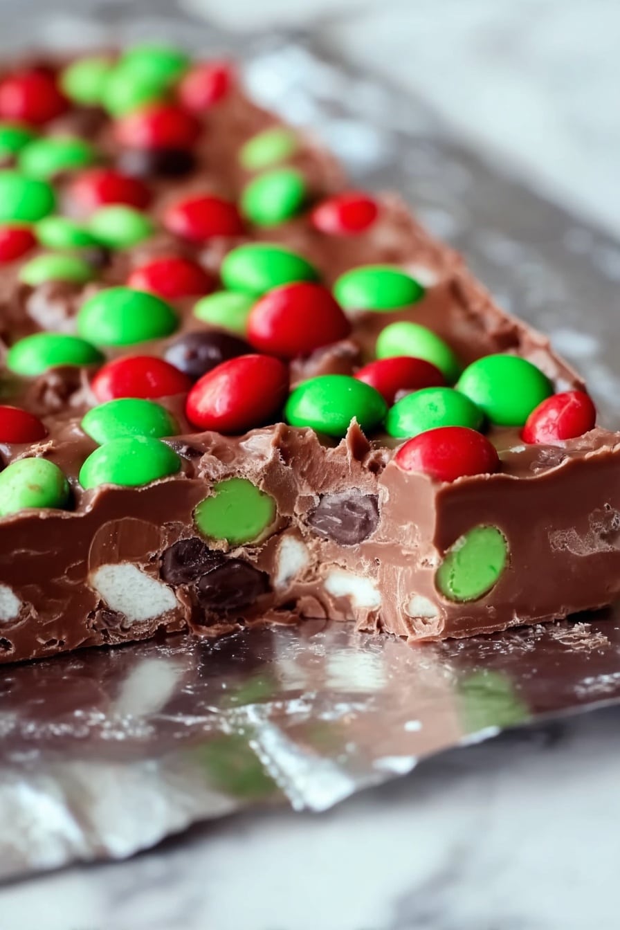The image shows a thick chocolate layer bar placed on a shiny silver foil with a piece removed from the front left side. The bar has three visible layers: the base layer is solid milk chocolate that holds the whole shape, the middle layer contains small white marshmallow bits embedded inside the chocolate, and the top layer is covered with bright red and green candy-coated chocolate pieces scattered evenly across the surface. The colors are vibrant, emphasizing the contrast between the dark brown chocolate and the candy's red and green. The background is a white marbled texture. Photo taken with an iphone --ar 2:3 --v 7 - M&M's Marshmallow Dream Bars, easy marshmallow dessert, no-bake chocolate bars, nostalgic candy treats, quick peanut butter bars