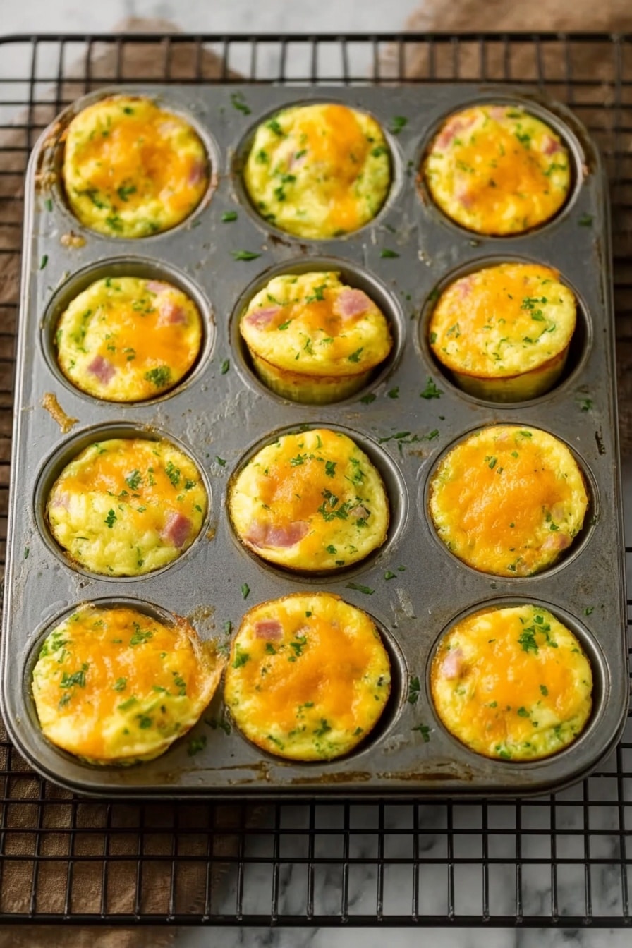 Ham and Cheese Egg Muffins Recipe 2 A metal muffin pan holds 12 small egg muffin cups, each filled with layers of fluffy yellow egg mixed with bits of green vegetables and pink ham. The muffins have a light golden top with melted orange cheese, creating a slightly bumpy and soft texture. Some muffins puff up more than others, showing a thick and airy inside with green herbs spread throughout. The pan rests on a rectangular black wire rack on a white marbled surface. Photo taken with an iphone --ar 2:3 --v 7 - Ham and Cheese Egg Muffins, Breakfast Egg Muffins, Easy Breakfast Recipes, Make-Ahead Egg Muffins, Protein-Packed Breakfast
