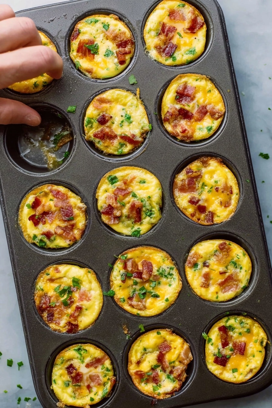 The image shows a dark metal muffin tray filled with twelve small egg bites. Each bite has a bright yellow egg base mixed with pieces of cooked bacon that have a reddish-brown color and small bits of green herbs or vegetables scattered on top and inside. The egg bites are slightly raised and have a soft, slightly shiny texture with some cheese melted and golden brown on top. The tray rests on a white marble surface, with a woman’s hand reaching toward the top left egg bite. Some small green herb pieces are scattered around the tray. photo taken with an iphone --ar 2:3 --v 7 - Bacon and Spinach Egg Muffins, breakfast egg muffins, healthy breakfast recipes, make-ahead breakfast ideas, easy brunch recipes