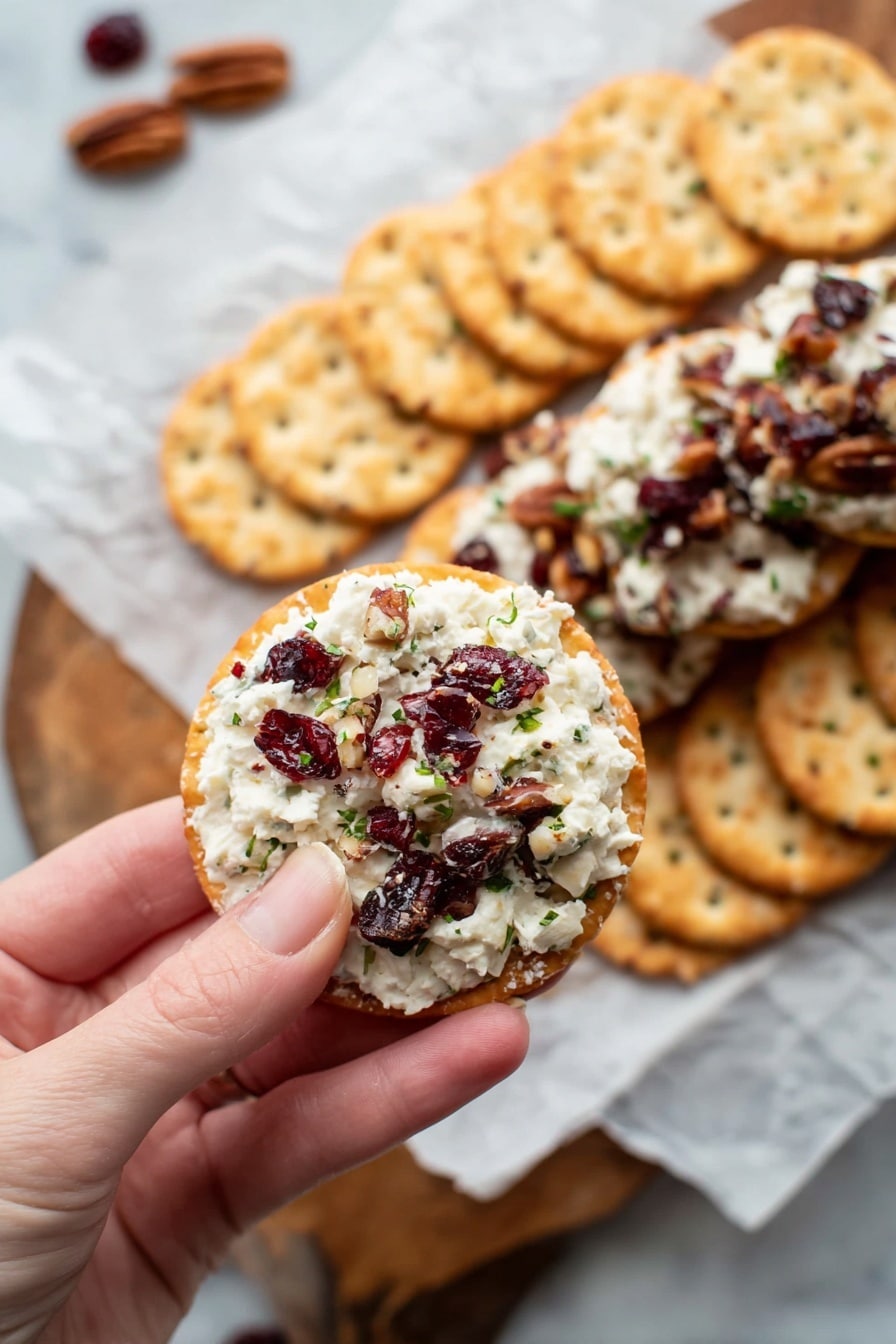 A log-shaped cheese ball covered with chopped nuts and pieces of dark red dried cranberries is shown close-up, resting on white parchment paper. The cheese inside is creamy white, mixed with bits of green herbs and pieces of cranberries and nuts. The outer surface of the log is rough with visible pieces of nuts and cranberries in warm brown, dark red, and green colors. Some crackers and scattered nut pieces appear blurred in the foreground on a white marbled surface. Photo taken with an iphone --ar 2:3 --v 7 - Cranberry Pecan Cheese Log, holiday cheese appetizer, festive cheese ball, easy party appetizers, creamy cheese and cranberry spread