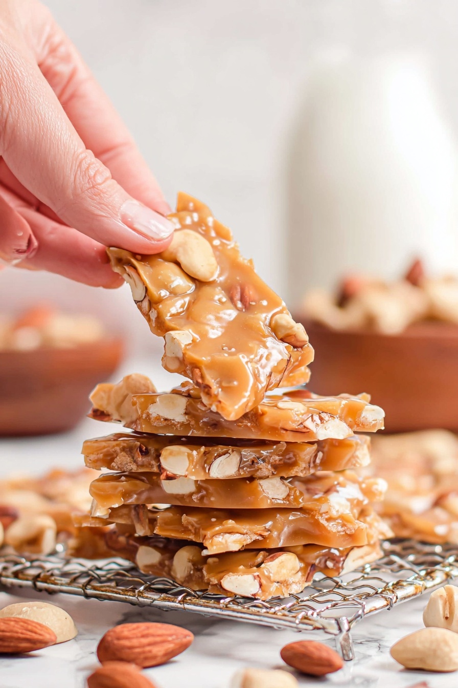 Homemade Nut Brittle Recipe 6 A woman's hand is holding up a piece of light brown nut brittle with visible whole almonds and cashews embedded in it. Below, there is a stack of four pieces of the same nut brittle, each piece thin and flat, showing off the nuts inside. The stack rests on a silver wire rack placed over a white marbled surface. Scattered around the rack are whole almonds and cashews, while the blurred background shows a white bottle and a wooden bowl. The overall image is bright and clear with warm tones. photo taken with an iphone --ar 2:3 --v 7 - Homemade Nut Brittle, nut brittle recipe, crunchy caramel brittle, homemade candy, nut brittle dessert
