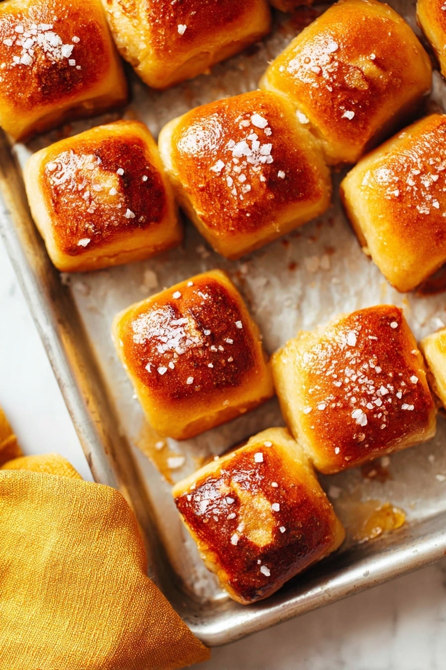 Soft Pretzel Bites Recipe 6 The image shows a metal baking tray holding about a dozen small square pretzel bites. Each piece has a golden brown top with a slightly darker toasted patch in the center and is sprinkled with coarse salt crystals. The background is a white marbled texture, and a mustard-yellow cloth is partially visible in the bottom left corner. The pretzel bites are neatly spaced, showing a soft and fluffy texture with a shiny crust. Photo taken with an iphone --ar 2:3 --v 7 - Soft Pretzel Bites, Soft Pretzel Bites Recipe, Chewy Pretzel Snacks, Baked Pretzel Bites, Easy Pretzel Bites