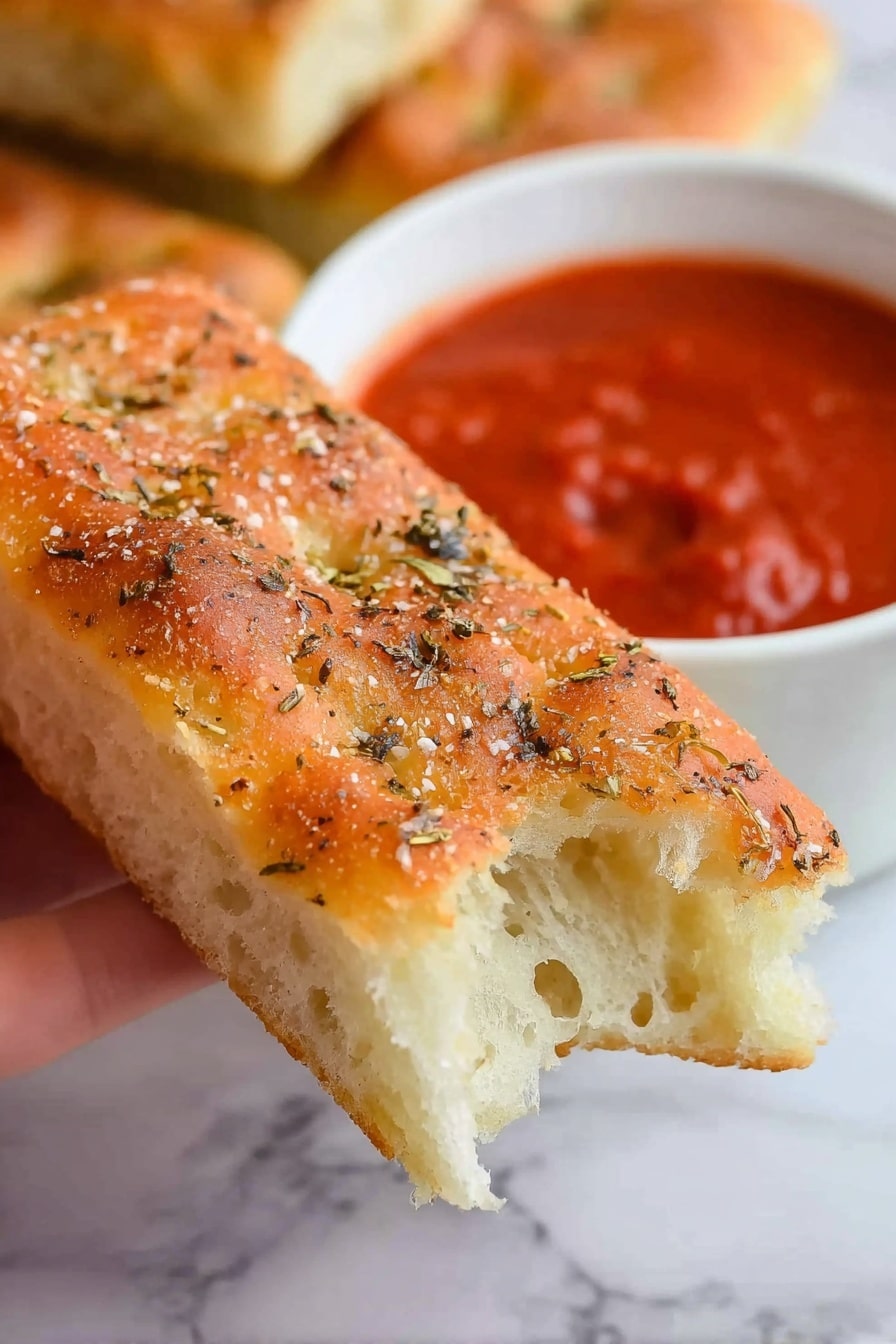 Copycat Pizza Hut Breadsticks Recipe 6 A close-up of a woman's hand holding a rectangular piece of focaccia bread with a light golden crust on top sprinkled with coarse salt and dried herbs, showing the soft, airy white inside at the bitten end. In the blurry background, a white bowl filled with thick red marinara sauce is visible, alongside another piece of focaccia bread partly visible on a white marbled surface. photo taken with an iphone --ar 2:3 --v 7 - Copycat Pizza Hut Breadsticks, homemade pizza breadsticks, soft buttery garlic breadsticks, how to make Pizza Hut breadsticks, best homemade breadsticks