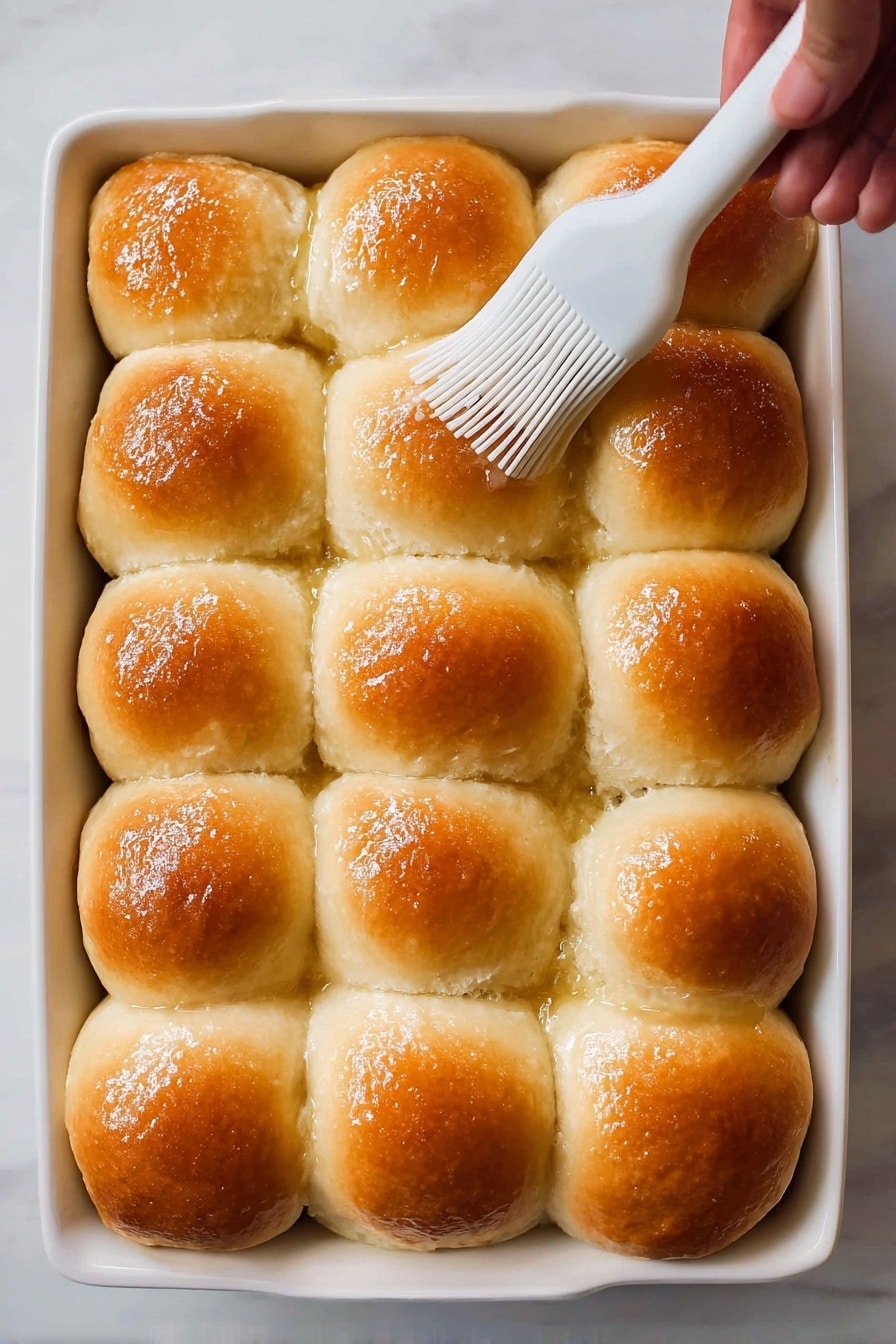A white baking dish holds fifteen soft, fluffy golden brown dinner rolls arranged in a 3 by 5 grid. Each roll has a shiny, smooth top with a slightly darker brown in the middle and a light fluffy texture on the sides where they touch. A white silicone brush held by a woman's hand gently spreads butter or glaze on the top of one roll near the center. The background is a white marbled texture. photo taken with an iphone --ar 2:3 --v 7 - Soft & Fluffy Dinner Rolls, dinner rolls recipe, easy dinner roll recipe, homemade dinner rolls, soft bread rolls